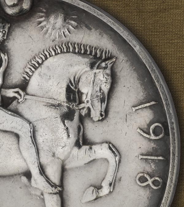 A detail, showing the head and front half of a horse, on the reverse of the 1914-18 War medal. The whole medal shows Saint George on horseback, armed with a short sword. The horse tramples on the Prussian eagle shield and the emblems of death, a skull and cross-bones. In the background are ocean waves and at the top the risen sun of Victory.