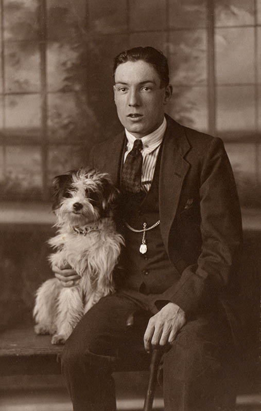 Studio portrait of William Pearson, 1919. He is sitting on a wooden bench holding a small, very hairy, dog in one hand and a walking stick in the other 